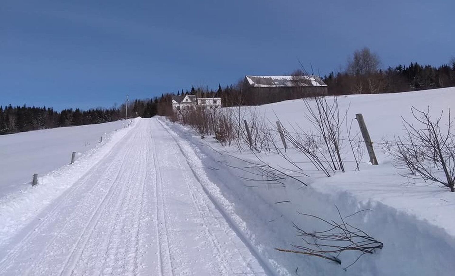 A snow-covered rural road with a barn and trees in the background.