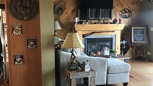A cozy living room with a fireplace, a sofa, a coffee table, and a wooden wall with clocks and signs. - WINTERFELL-NH BEST OF NH GORGEOUS LAKE &  MOUNTAIN VIEWS,  NEVER PP ADDS (Pittsburg)