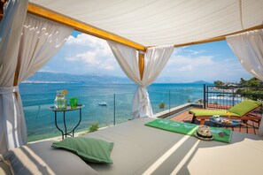 Ein Balkon mit einem Bett, einem Tisch mit Getränken und Blick auf Meer und Berge.