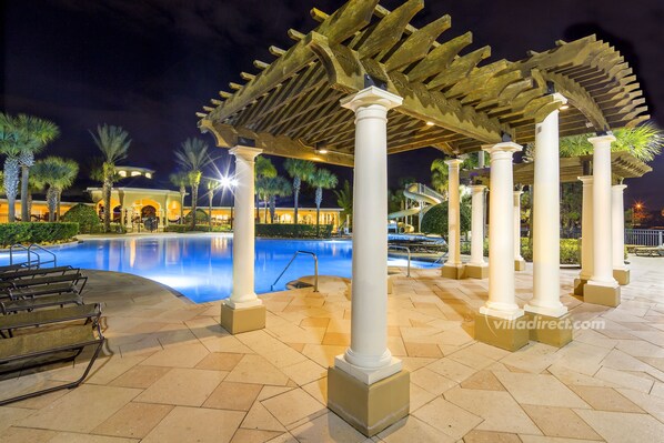 A nighttime pool area with a pergola, palm trees, and a pool with a clear blue water surface.