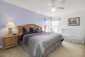 Quarto com cama, mesas de cabeceira, ventoinha no teto, janela com cortinas e berço.