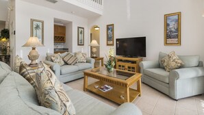 A spacious living room with two sofas, a coffee table, and a television. There are framed pictures on the walls and a balcony above.