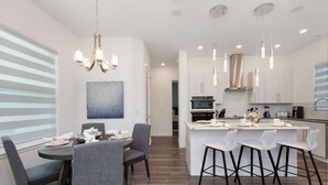 A modern kitchen with a central island, pendant lighting, and a dining area with grey chairs. - Paradise at Providence by VillaDirect (Davenport)