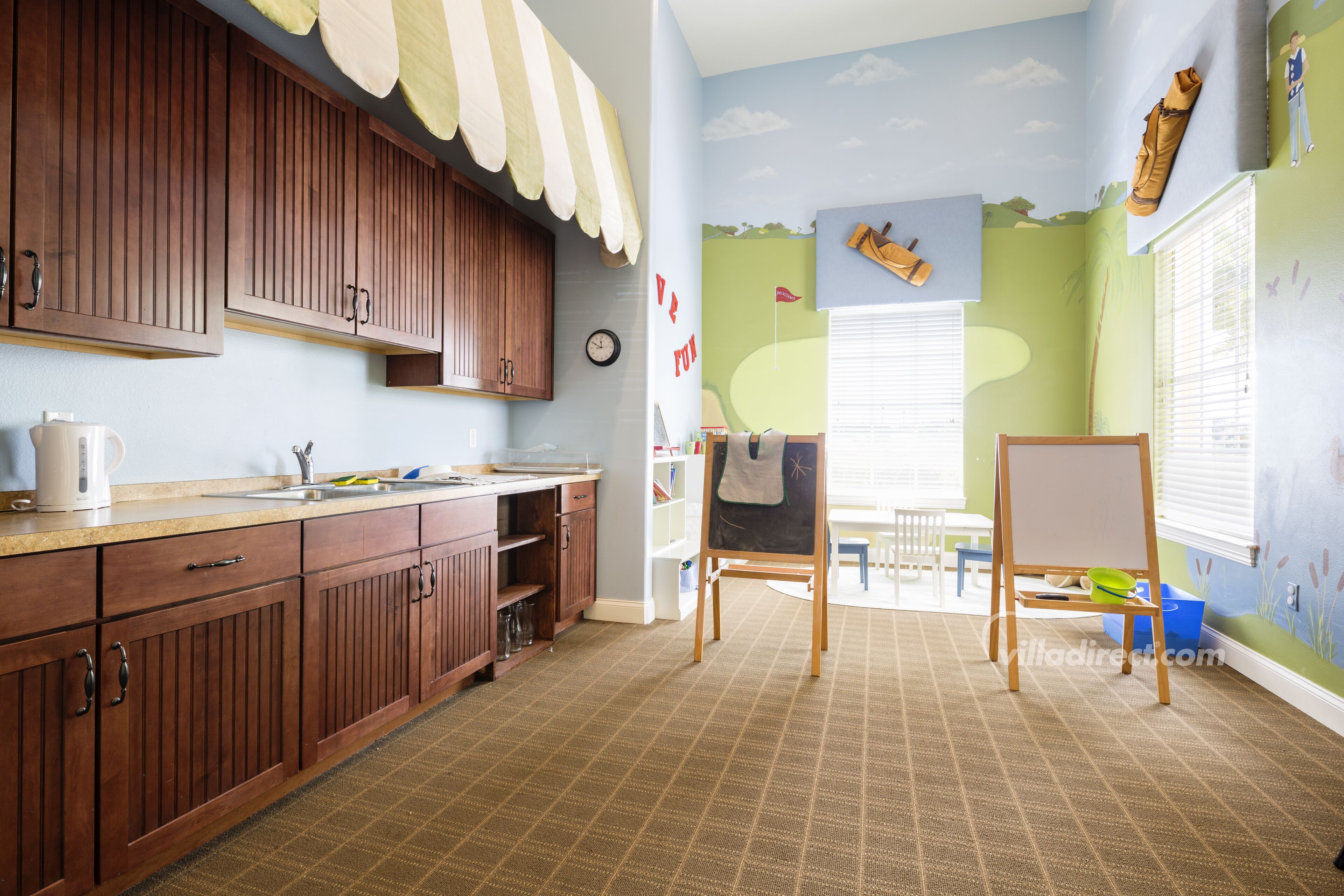 A kitchen with wooden cabinets, a sink, and a chalkboard. There is a window with blinds and a small table with a chair.