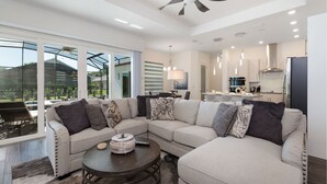 A modern living room with a sectional sofa, a coffee table, and a dining area in the background. - Paradise at Providence by VillaDirect (Davenport)
