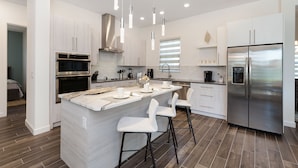 A modern kitchen with white cabinetry, stainless steel appliances, and a central island with bar stools. - Paradise at Providence by VillaDirect (Davenport)