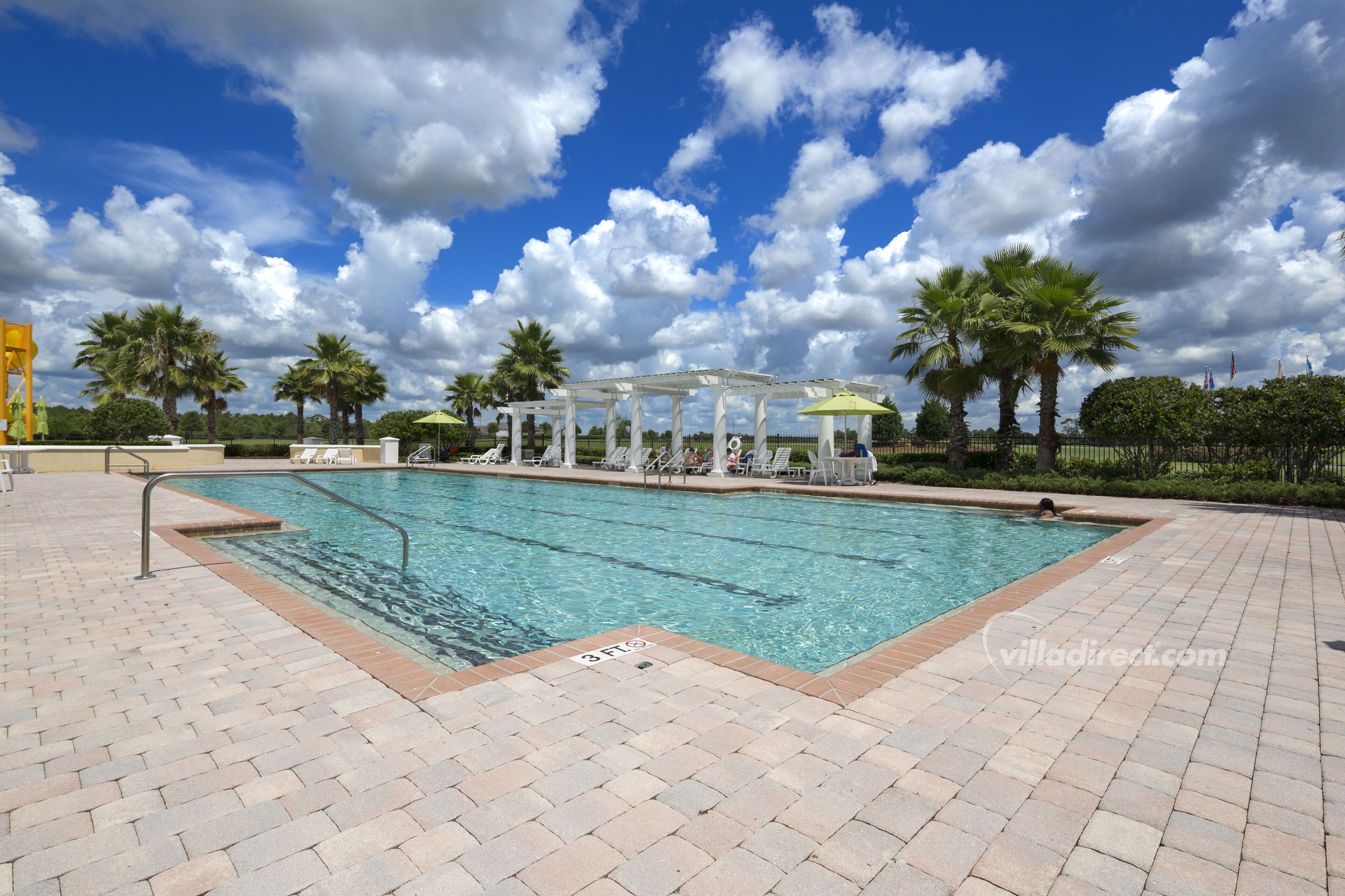 A swimming pool with a white pergola, palm trees, and a clear blue sky.