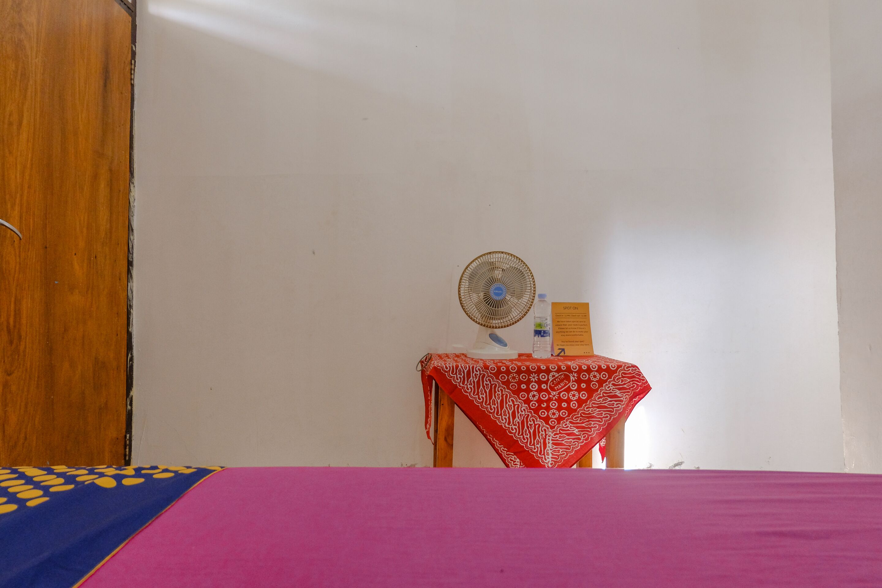Una habitación con una cama, una puerta de madera, una mesa con mantel rojo y un ventilador de pie.