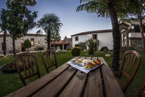 An outdoor dining area with a wooden table set for breakfast, surrounded by stone buildings and palm trees.