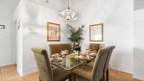 Un comedor con una mesa de vidrio, cuatro sillas tapizadas, una araña, obras de arte enmarcadas y una planta en maceta.