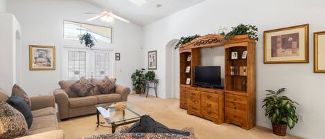 A spacious living room with a high ceiling, a wooden entertainment center, and a large window providing natural light.