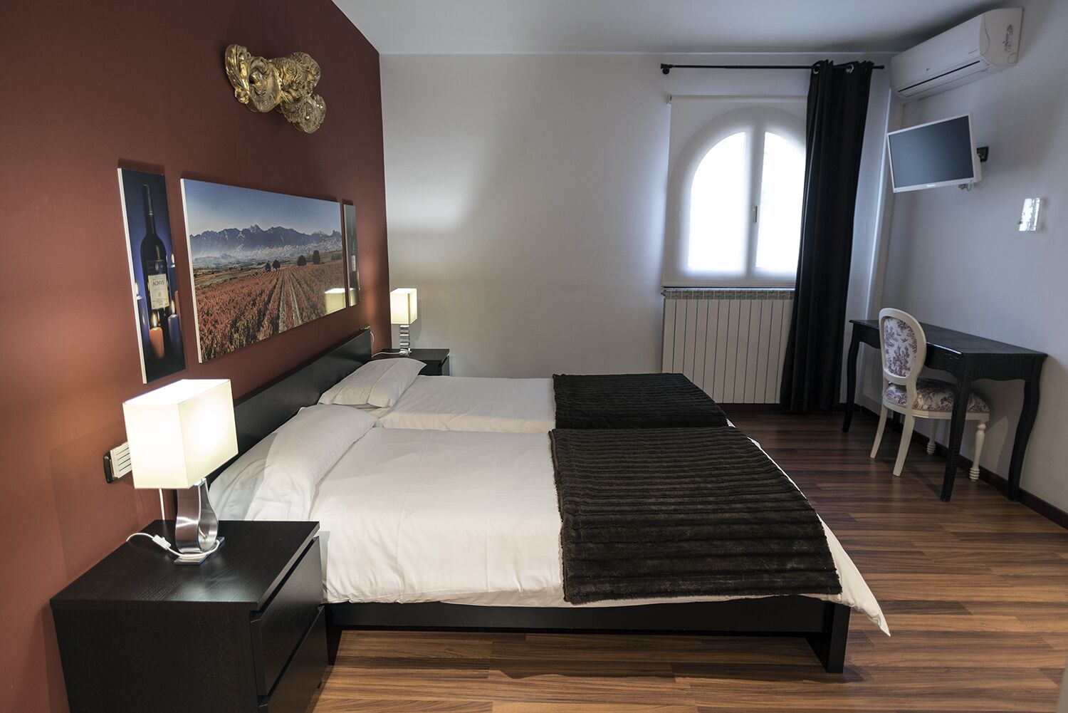 A hotel room with a double bed, bedside tables with lamps, a desk with a chair, a TV, and a large window with curtains.