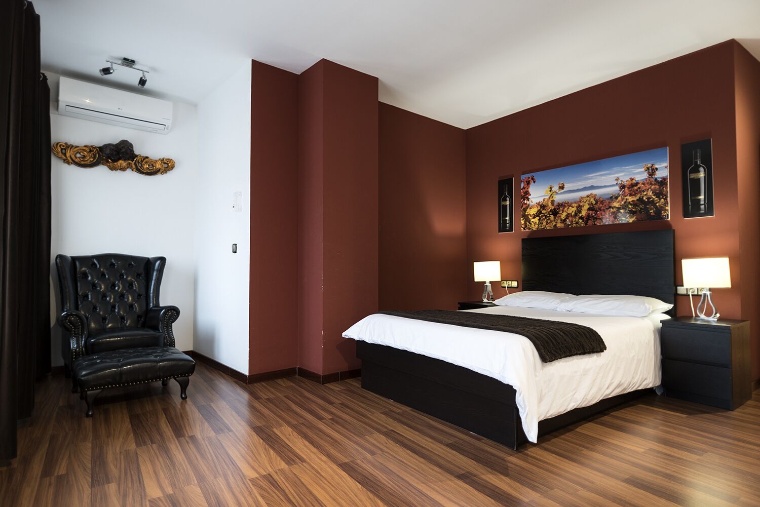 A hotel room with a large bed, a chair, a painting, and a wall-mounted air conditioner.