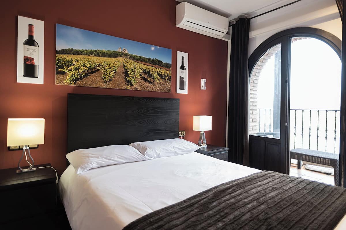 A hotel room with a large bed, two bedside tables with lamps, a wall-mounted air conditioner, and a large window with a balcony.