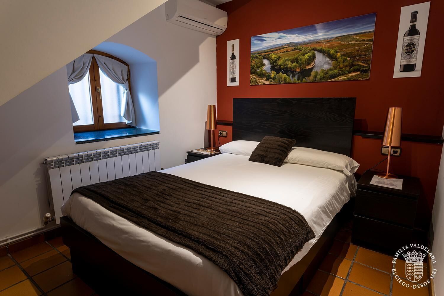 A bedroom with a large bed, a window with curtains, a radiator, and artwork on the walls.