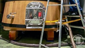 Iemand op een ladder die aan een groot houten vat werkt, met meetinstrumenten en kleppen, vermoedelijk in een opslag- of verwerkingsruimte.