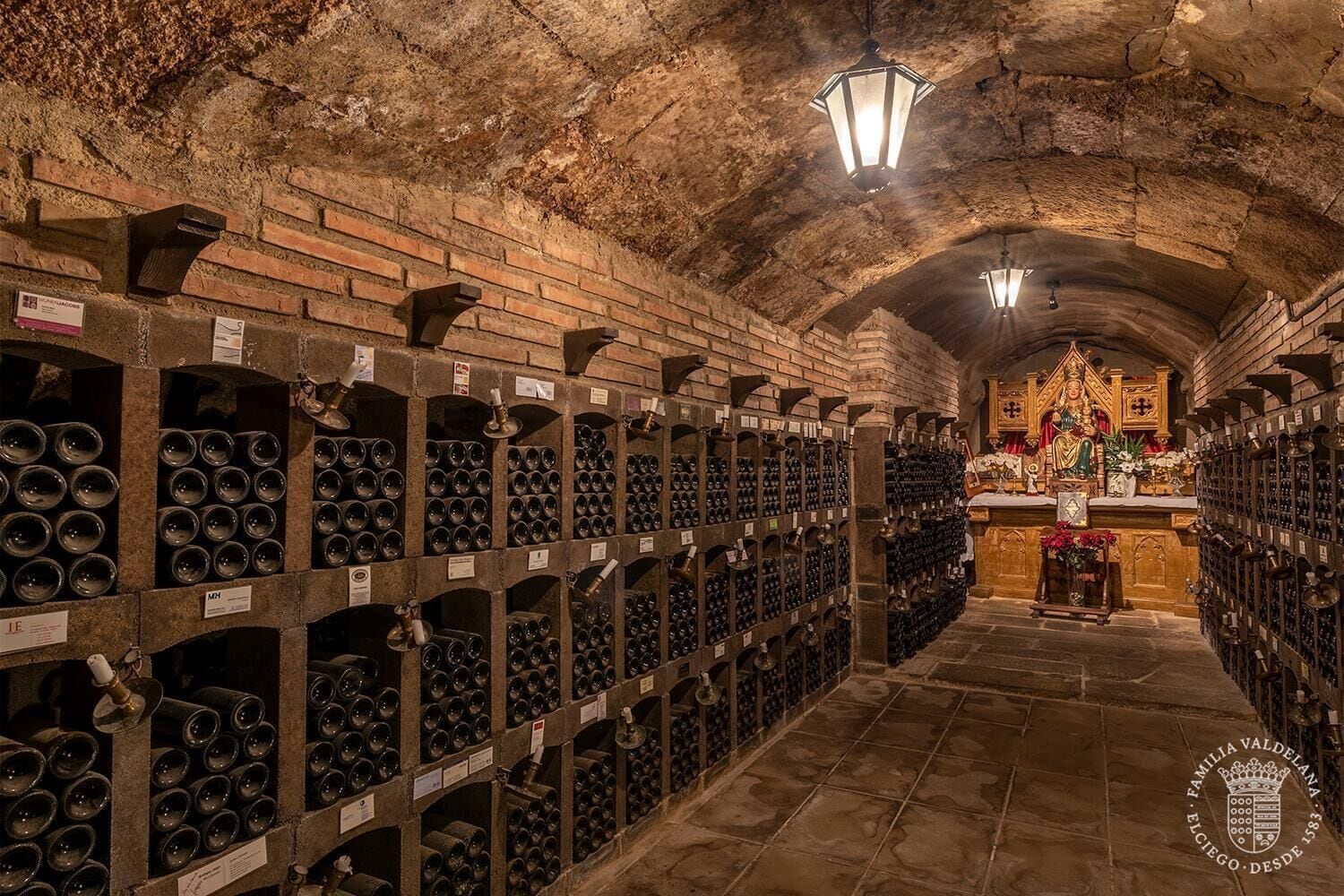Una bodega con hileras de botellas de vino en estantes, techo de piedra y una estatua al fondo.