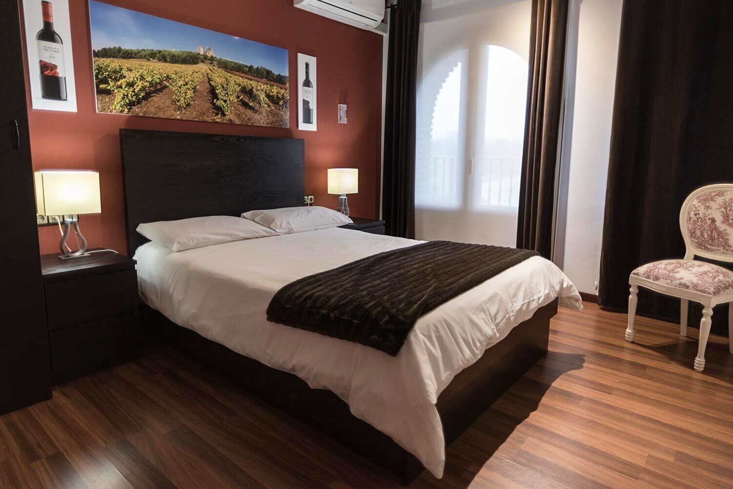 Habitación de hotel con una cama grande, dos mesitas de noche con lámparas, una silla y una pared decorada con cuadros de viñedos y botellas de vino.