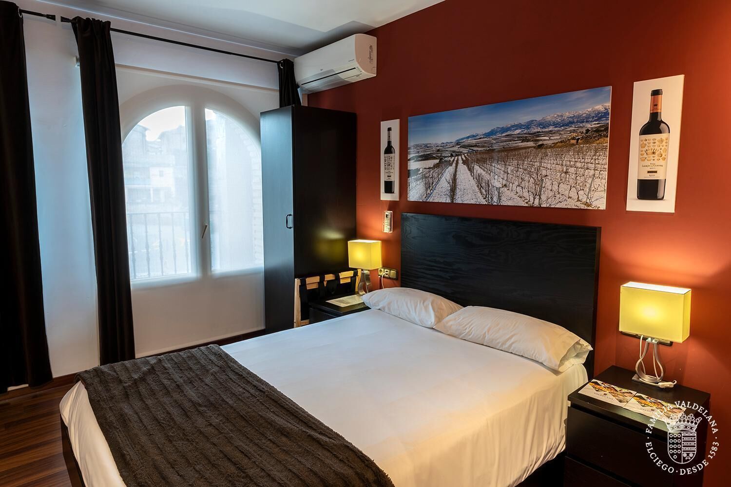 A hotel room with a large bed, two bedside tables with lamps, a wall-mounted air conditioner, and a scenic vineyard painting.