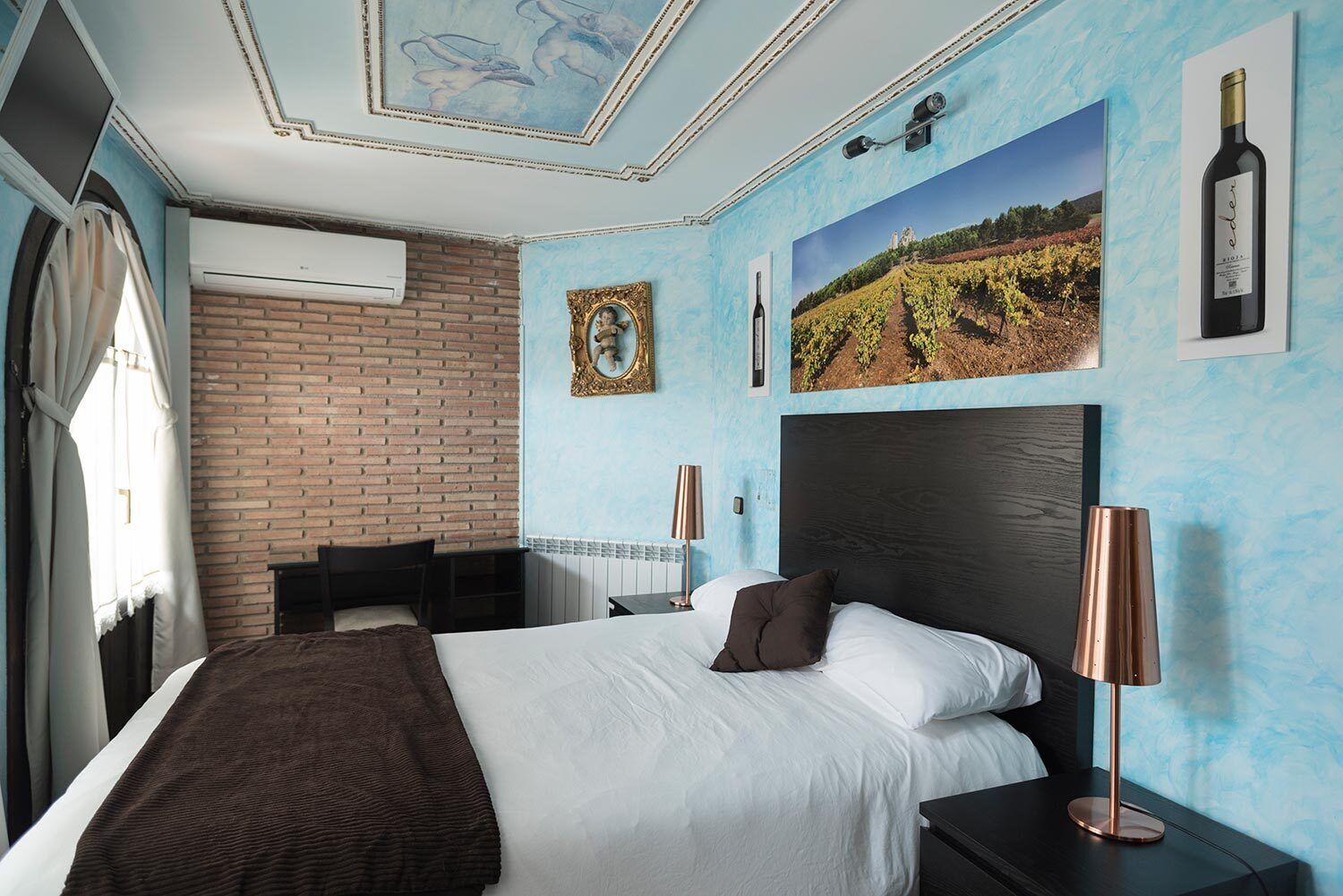 Een hotelkamer met een bed, een bureau, een stoel, een lamp en een afbeelding van een wijngaard aan de muur.