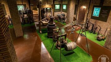 Una exposición de museo con artefactos históricos y maquinaria, incluyendo una gran rueda de madera y diversas herramientas, sobre fondos de paredes de ladrillo y suelo verde.