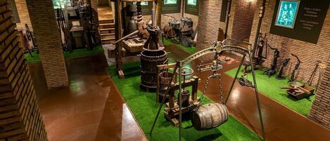 A museum exhibit with historical artifacts and machinery, including a large wooden wheel and various tools, set against brick walls and green flooring.