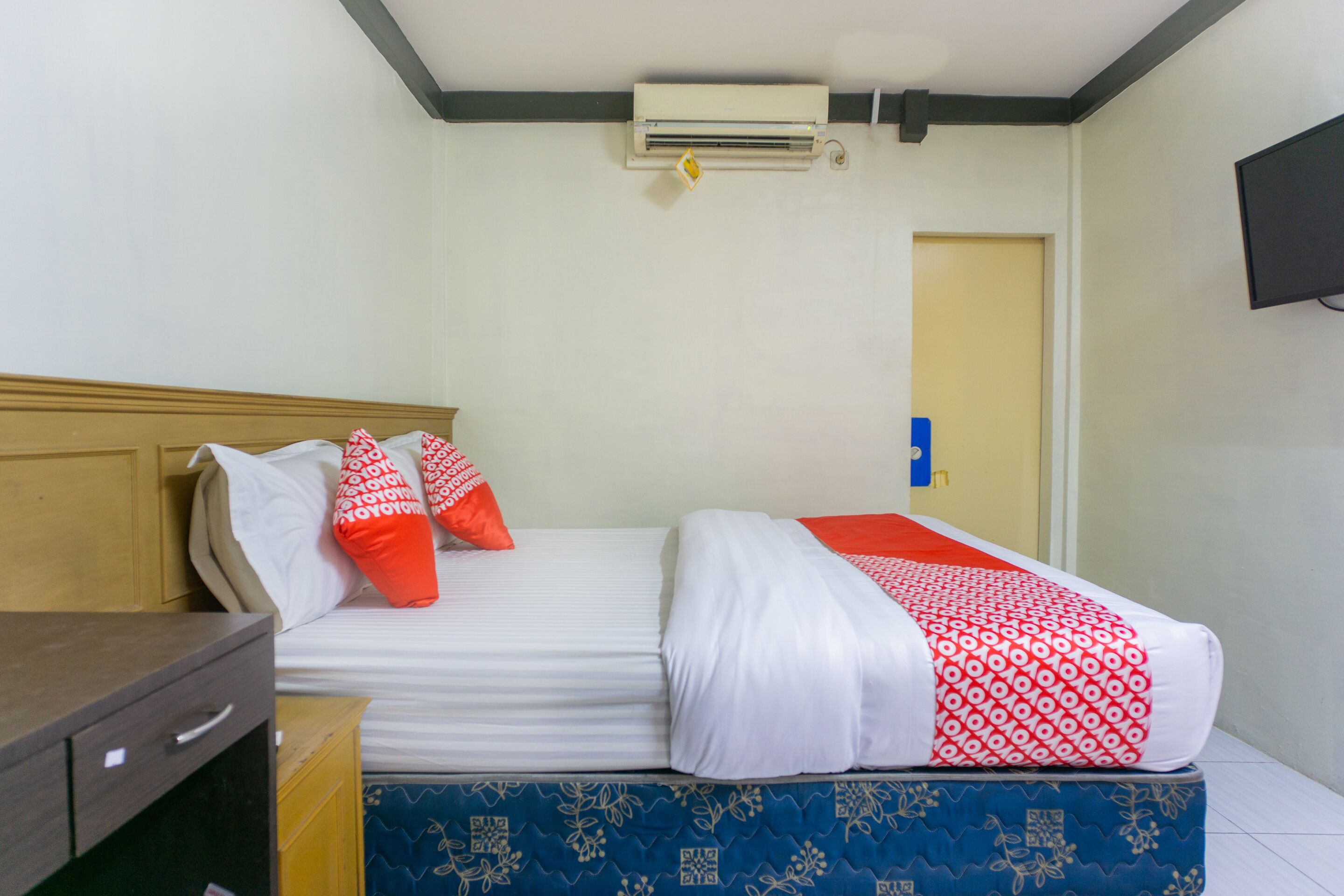 A hotel room with a bed, a wooden nightstand, a desk, an air conditioning unit, and a television on the wall.