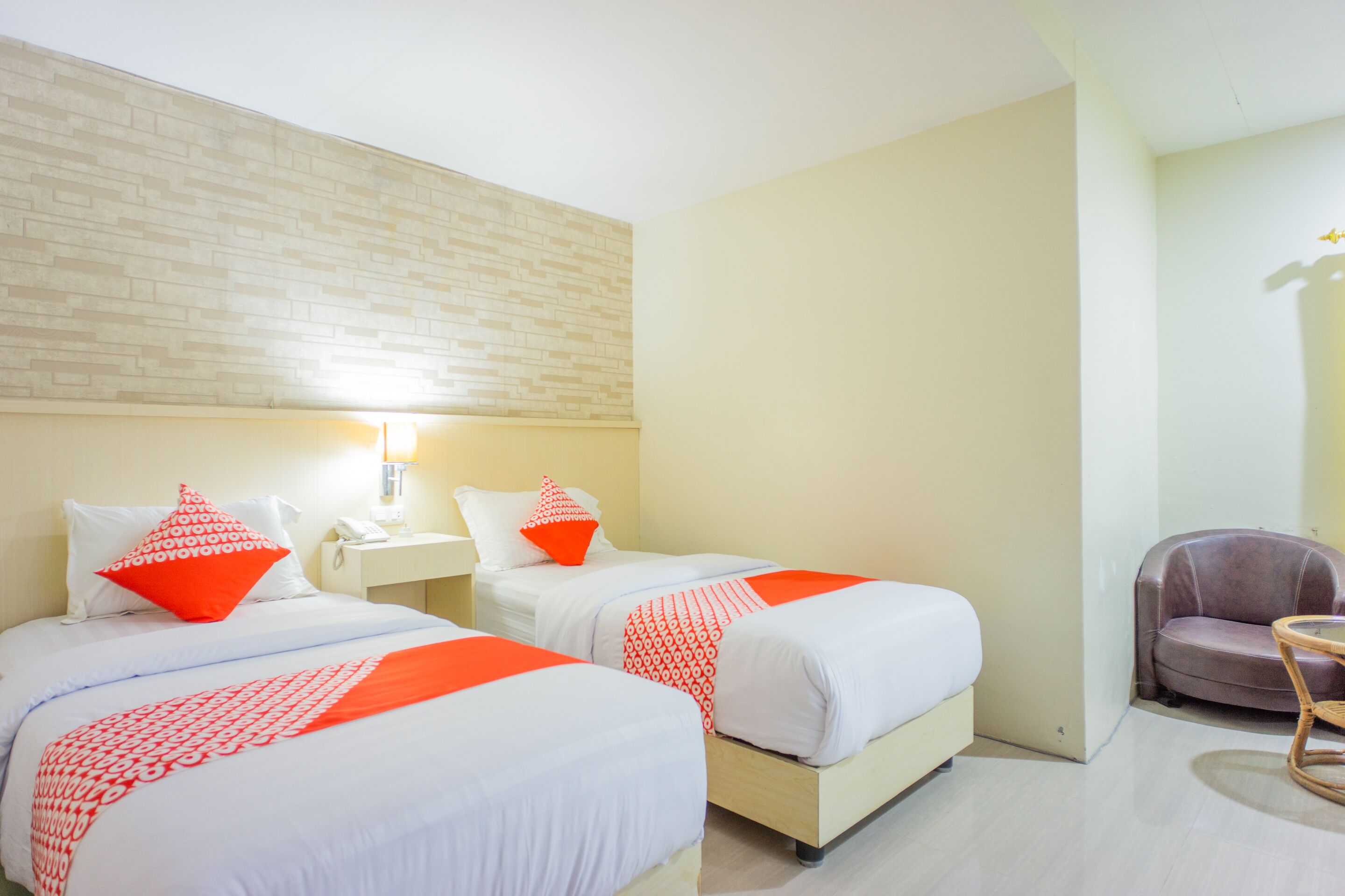 Quarto de hotel com duas camas, uma mesa pequena, uma cadeira e parede com padrão de tijolos.