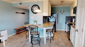 A compact kitchen with a round dining table, two chairs, a bench, a clock, and a refrigerator. - Walk to Sandfest, Shared Pools, Boardwalk to the Beach (Port Aransas)