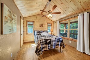 Une chambre avec un plafond en bois, un lit recouvert d’une couverture à carreaux, deux fenêtres et un ventilateur de plafond.