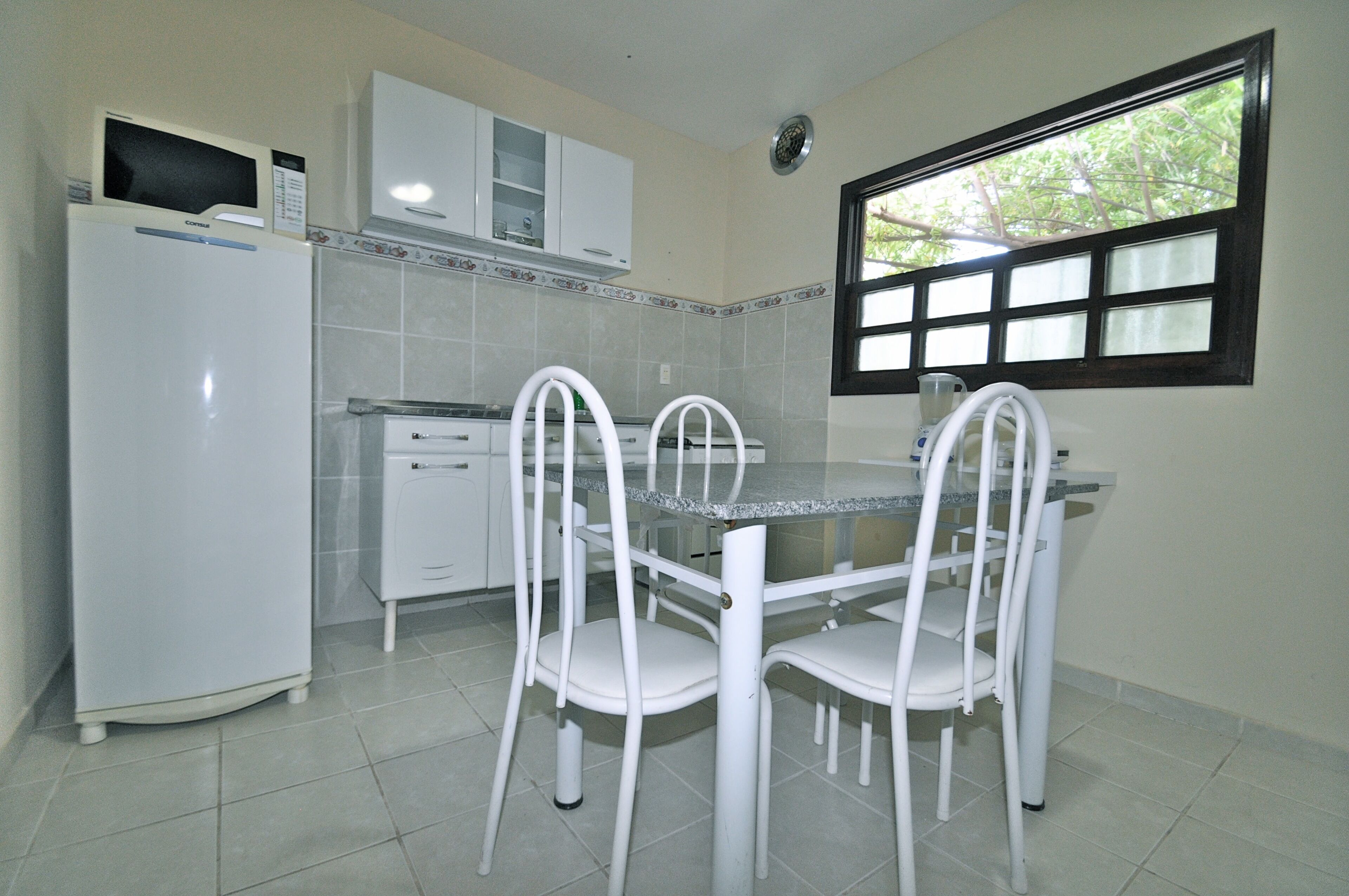 Uma cozinha compacta com eletrodomésticos brancos, painel de azulejos e janela com vista para árvores.