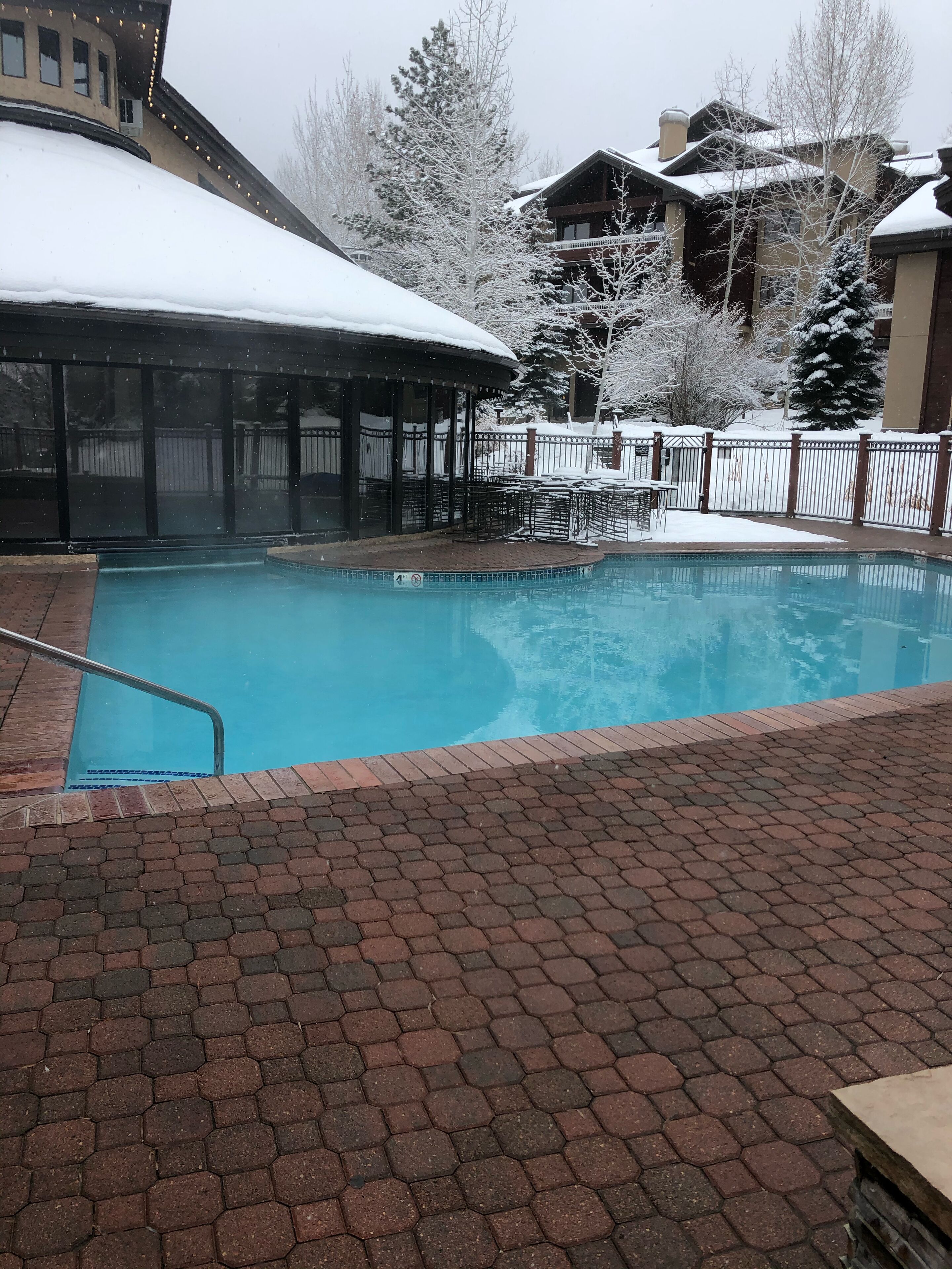 A swimming pool with a brick deck, surrounded by a snow-covered fence and buildings.