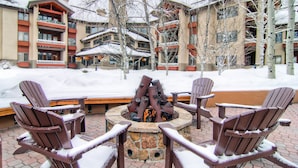 Un área de estar al aire libre cubierta de nieve con sillas Adirondack y un pozo de fuego de piedra.