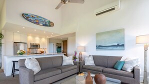 A modern living room with a grey sofa, a glass coffee table, and a kitchen in the background. - Ekahi 34F - Best Panoramic Ocean View!! (Kihei)