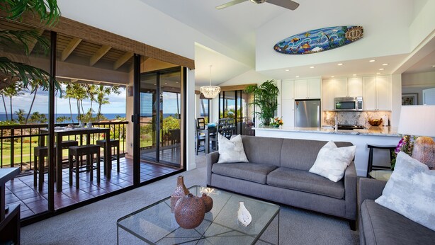 A modern living room with a glass coffee table, a grey sofa, and a dining area with a view of the ocean. - Ekahi 34F - Best Panoramic Ocean View!! (Kihei)