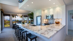 A modern kitchen with a large island, bar stools, and a view of the ocean. - Ekahi 34F - Best Panoramic Ocean View!! (Kihei)