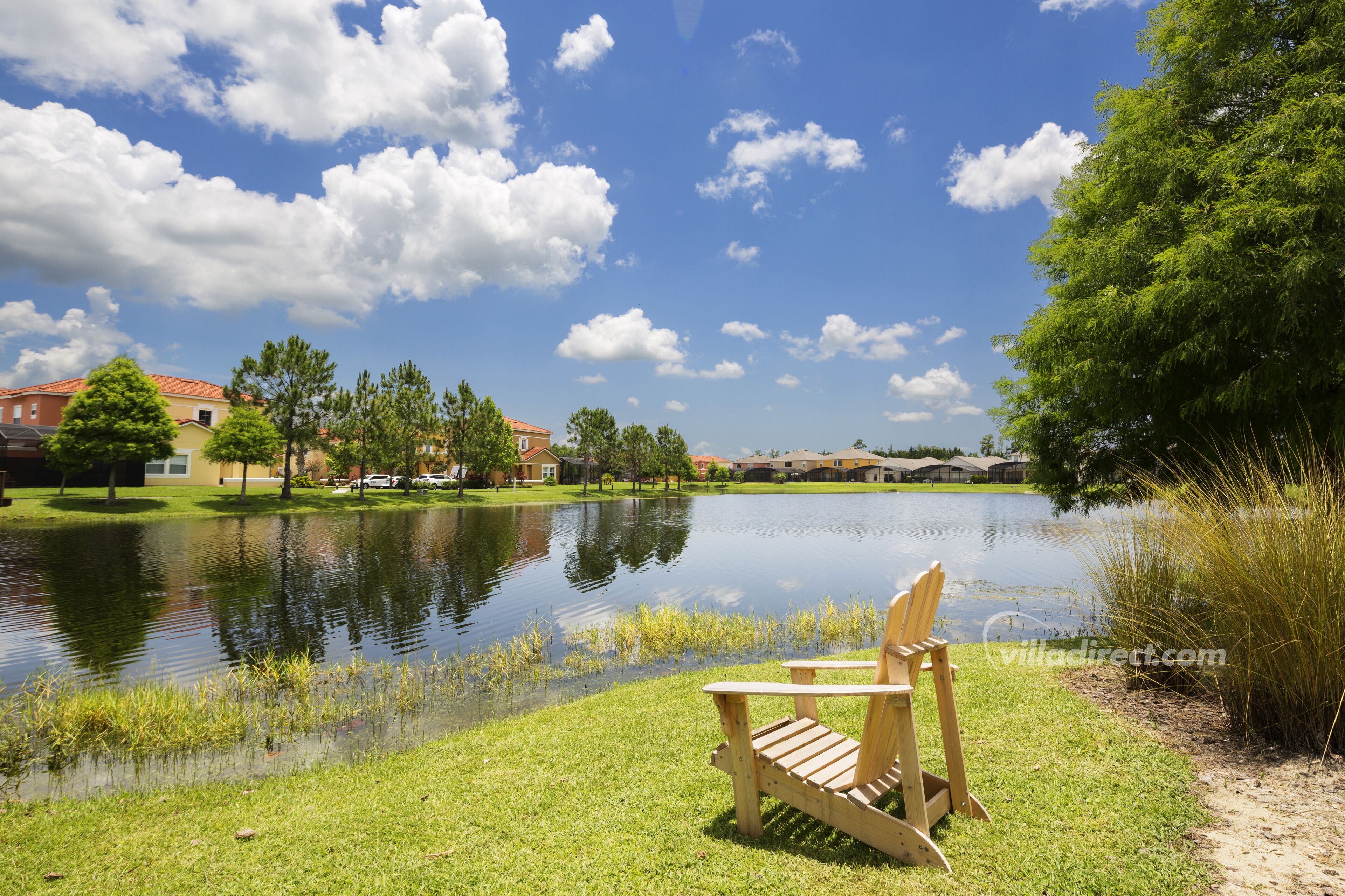 A lakeside view with a wooden chair by the water, houses in the background, and a clear sky.
