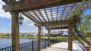 Une pergola en bois, dotée d’une rambarde en métal, surplombe un lac, et est entourée d’immeubles et de verdure.