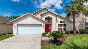 Une maison à deux étages avec une porte rouge, un garage et un petit jardin devant, où se trouve un palmier.