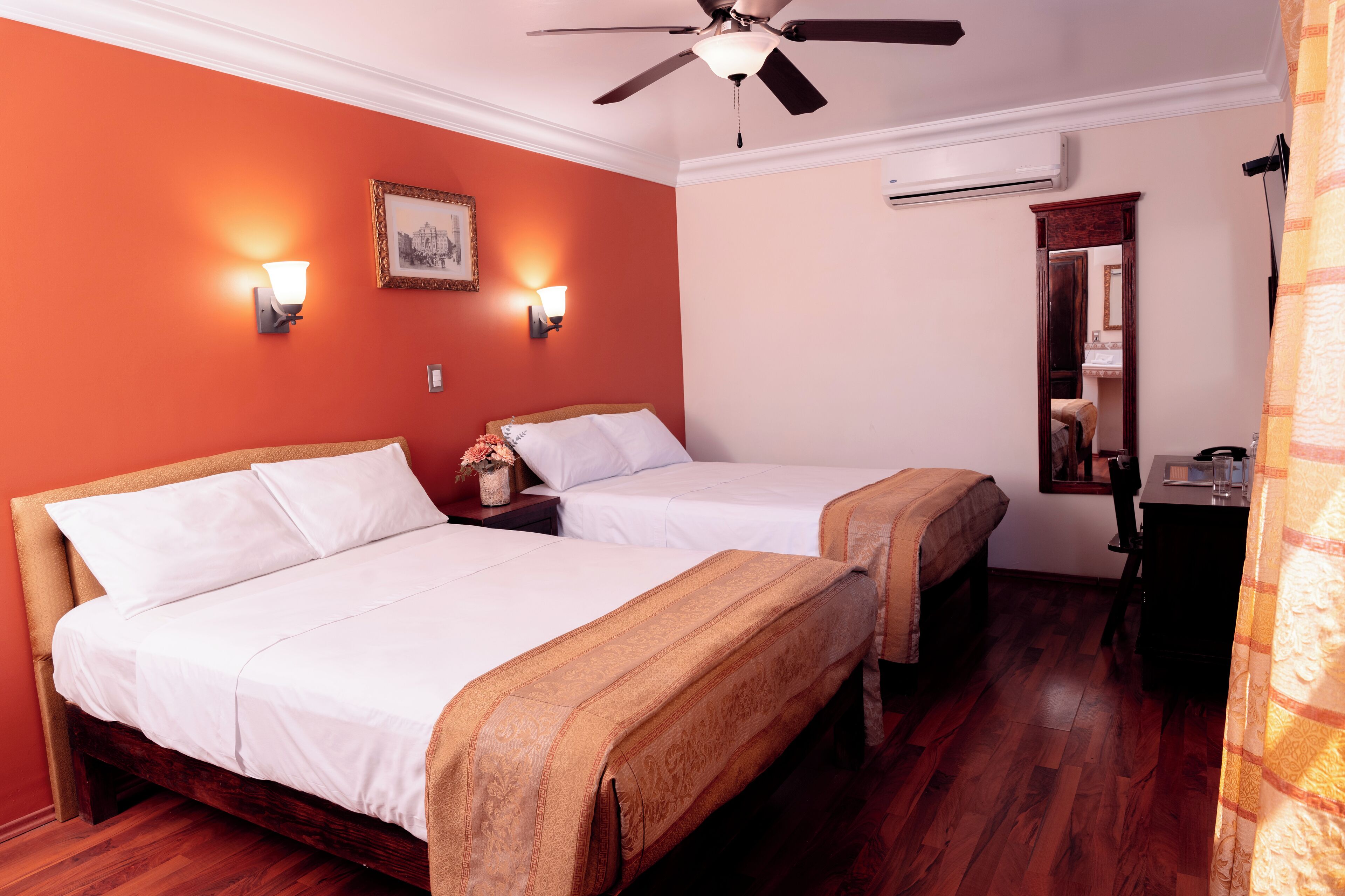 A hotel room with two beds, a ceiling fan, wall-mounted lights, a mirror, and a small desk.