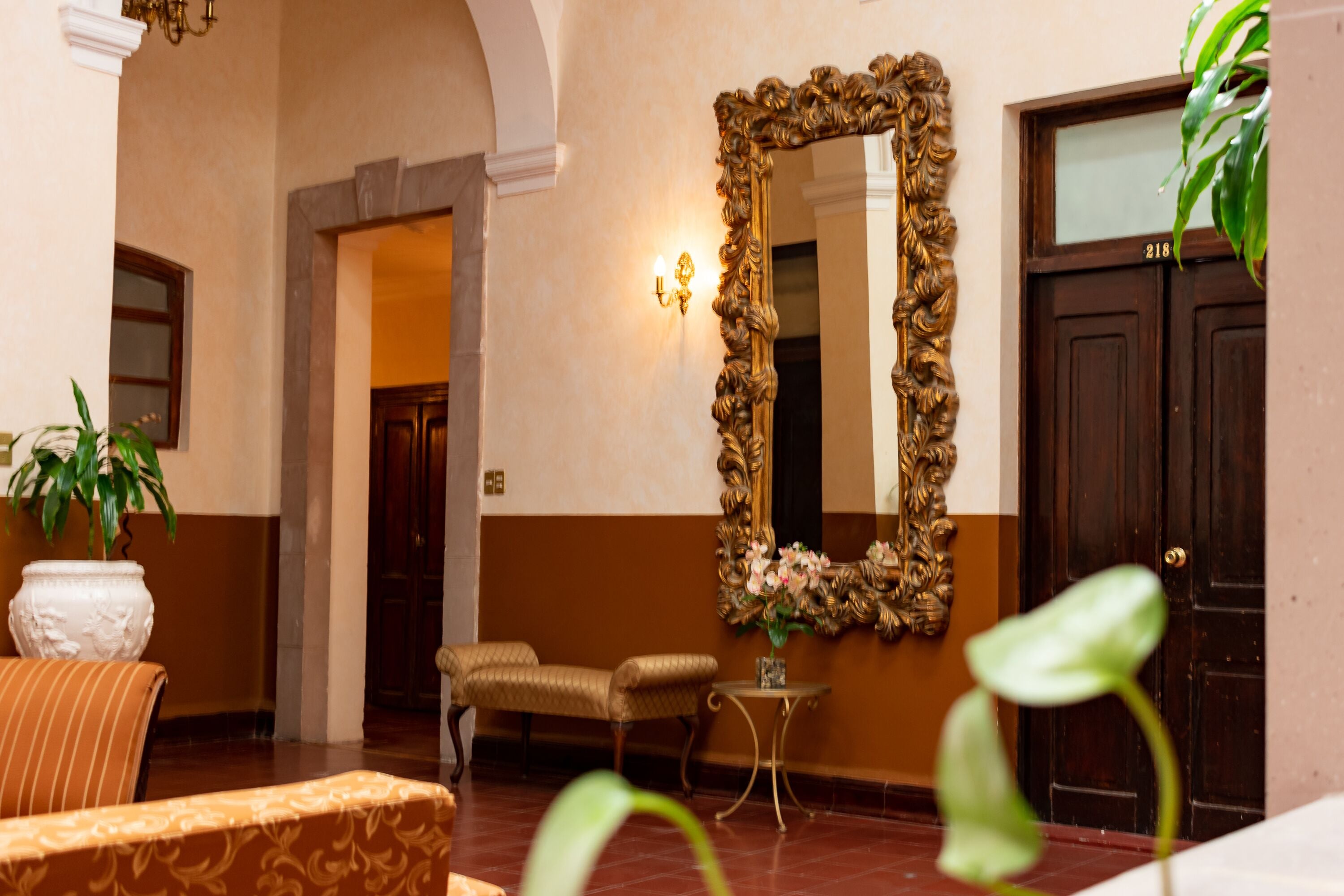 A lobby with a large ornate mirror, a sofa, a chair, a small table with a vase, and a potted plant.
