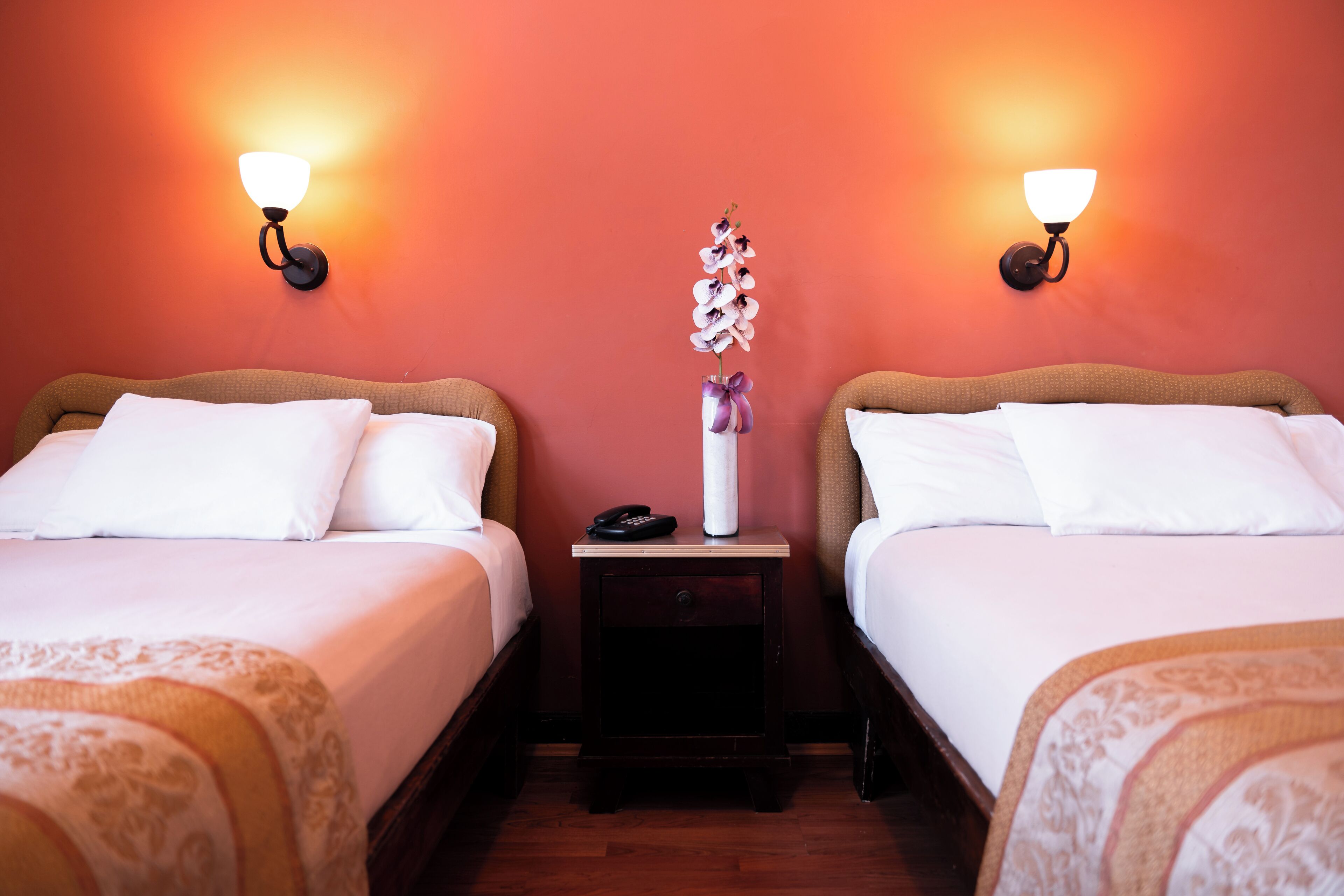 A hotel room with two beds, a nightstand with a vase of flowers, and wall-mounted lamps.