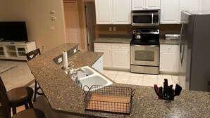 A kitchen with white cabinets, a microwave, stove, and refrigerator. There is a granite countertop, a sink, and a small kitchen island with a cutting board and knife block.