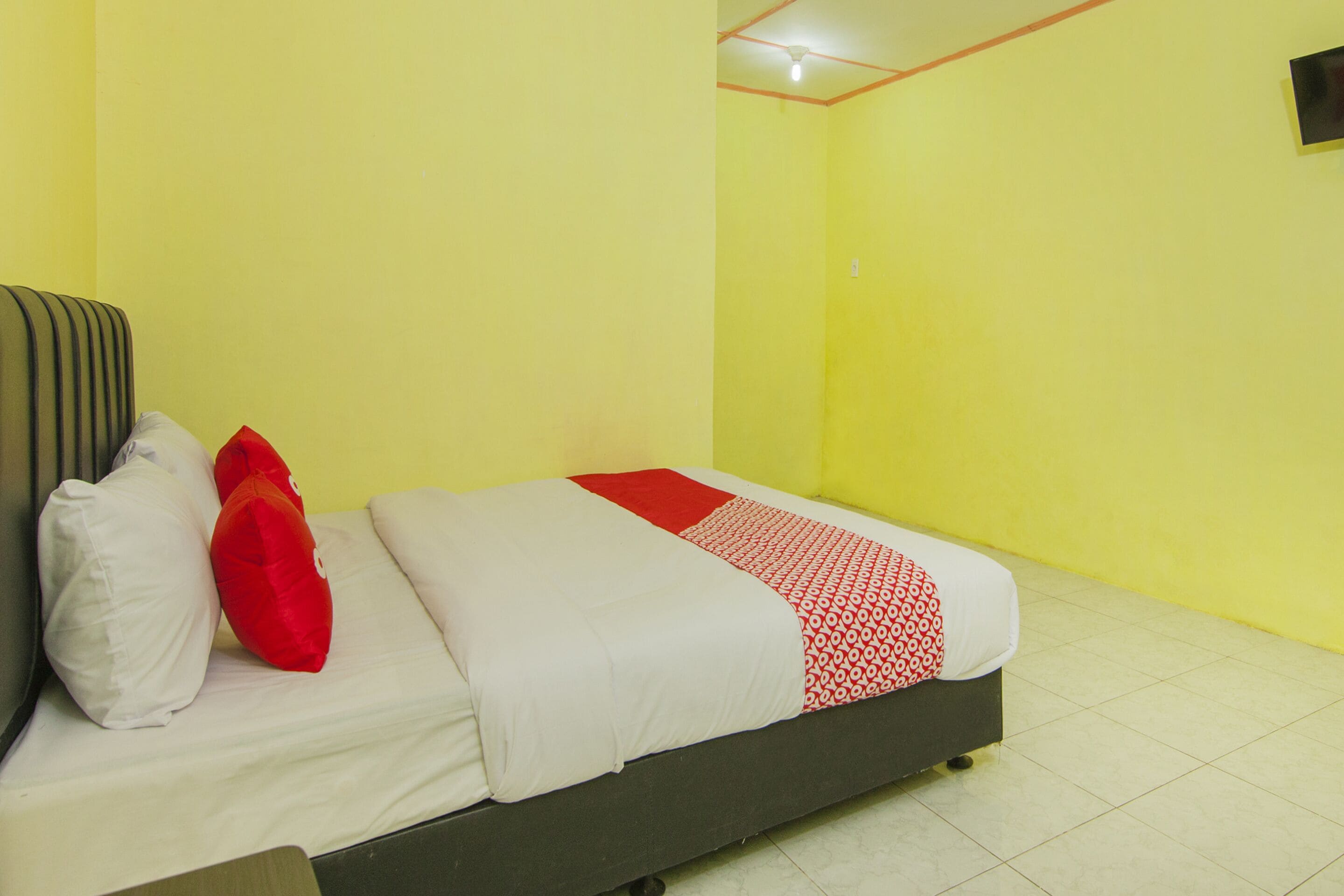 Una cama con ropa de cama blanca y roja, una almohada roja y una almohada blanca en una habitación con paredes amarillas y suelo de baldosas.