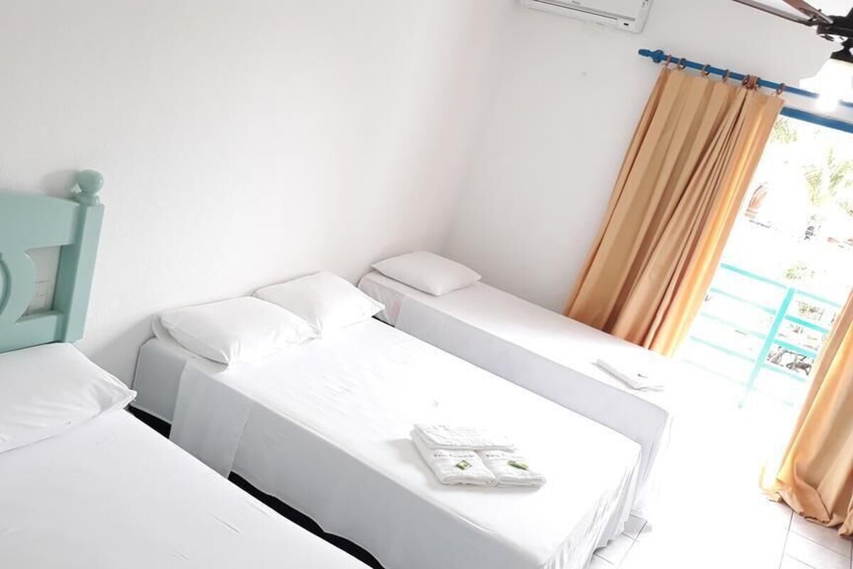 A room with two single beds, each with white bedding and a teal headboard. There is a window with beige curtains, and a small table with a phone and a book.