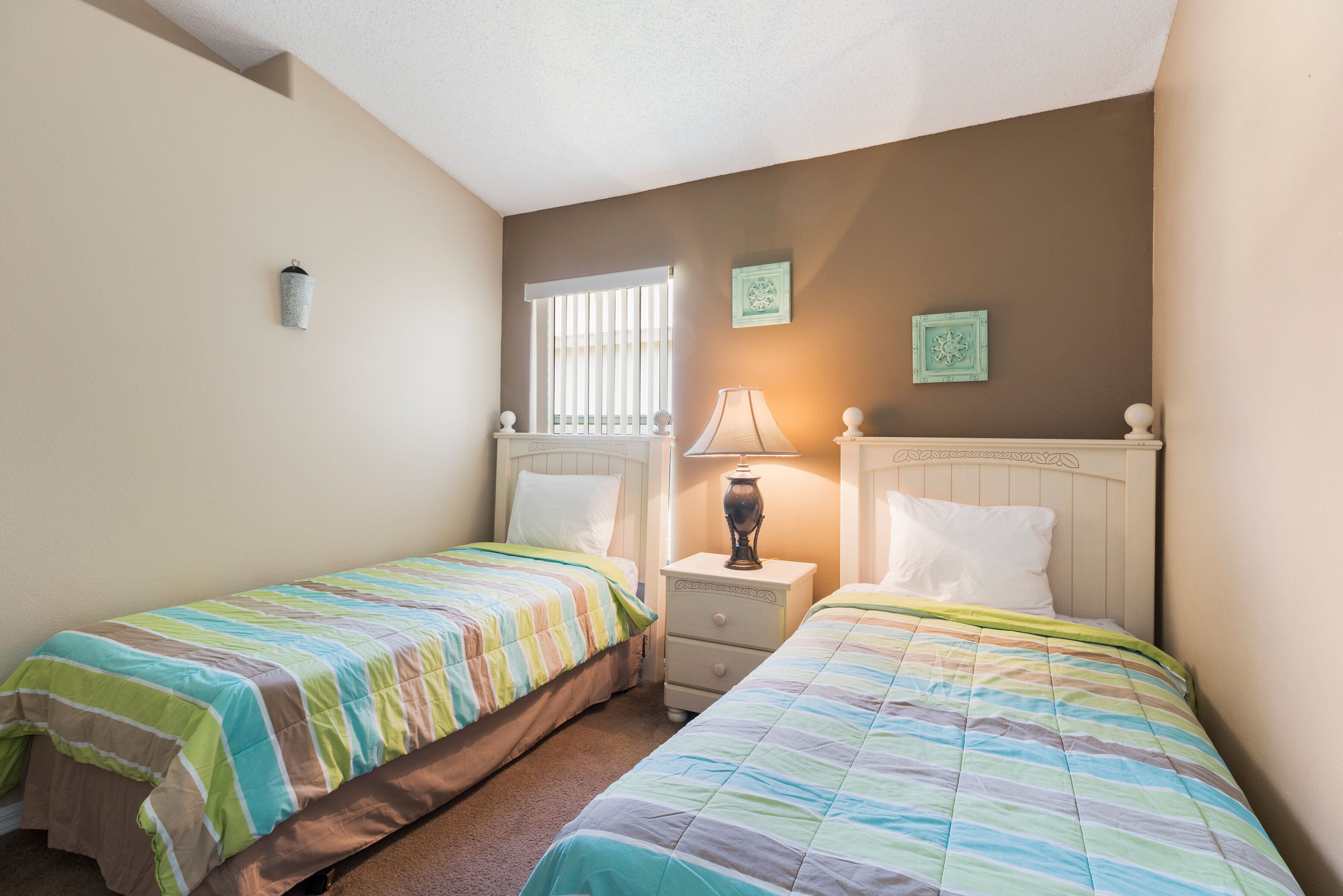 A bedroom with two beds, a nightstand with a lamp, a window with blinds, and two wall-mounted picture frames.