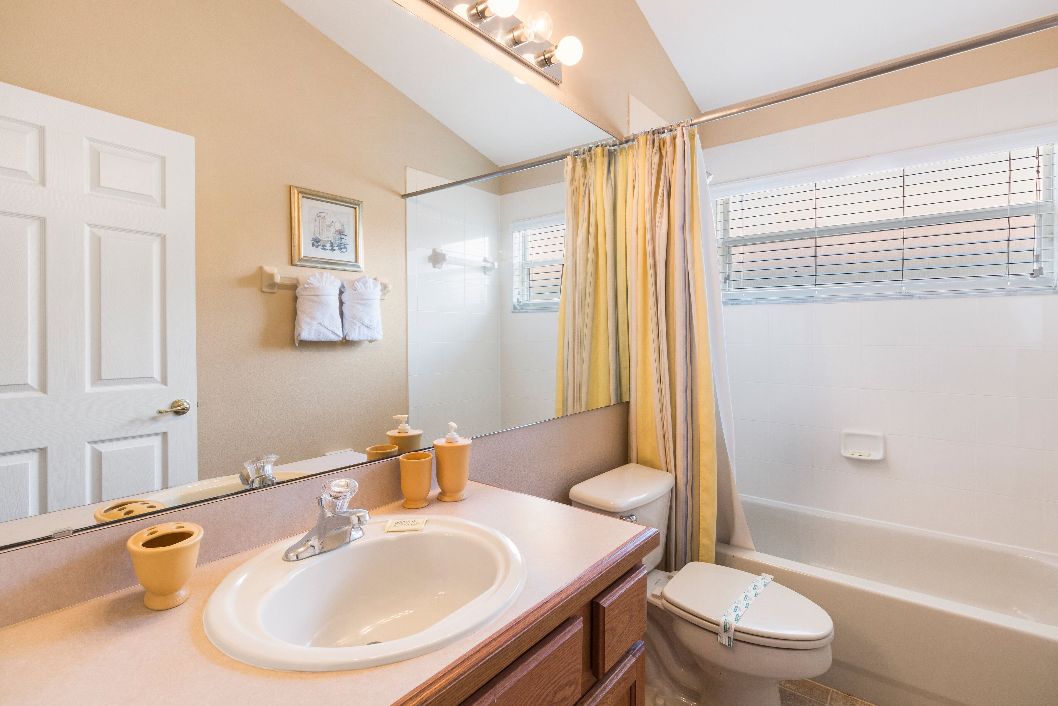 A bathroom with a white sink, mirror, and toilet. There is a shower curtain and a window with blinds.