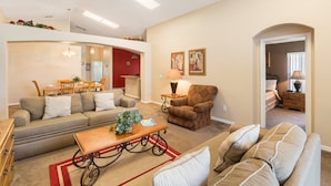 A spacious living room with a sofa, armchair, and coffee table. There is a dining area with a table and chairs, and a hallway leading to another room. - Sunshine Villa by VillaDirect (Kissimmee)