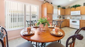 A dining area with a wooden table, chairs, and a vase with flowers. There is a window with blinds, a kitchen with wooden cabinets, and a microwave. - Sunshine Villa by VillaDirect (Kissimmee)