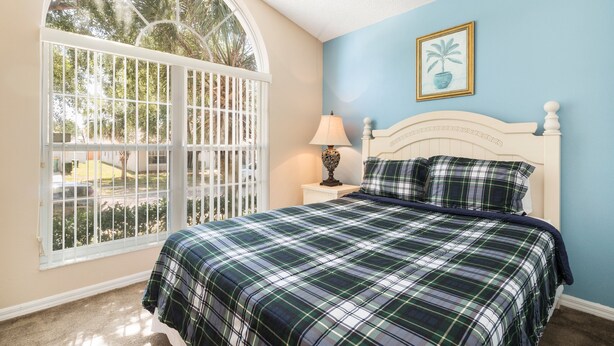 A bedroom with a large window, a bed with a plaid bedspread, a nightstand with a lamp, and a framed picture on the wall. - Sunshine Villa by VillaDirect (Kissimmee)